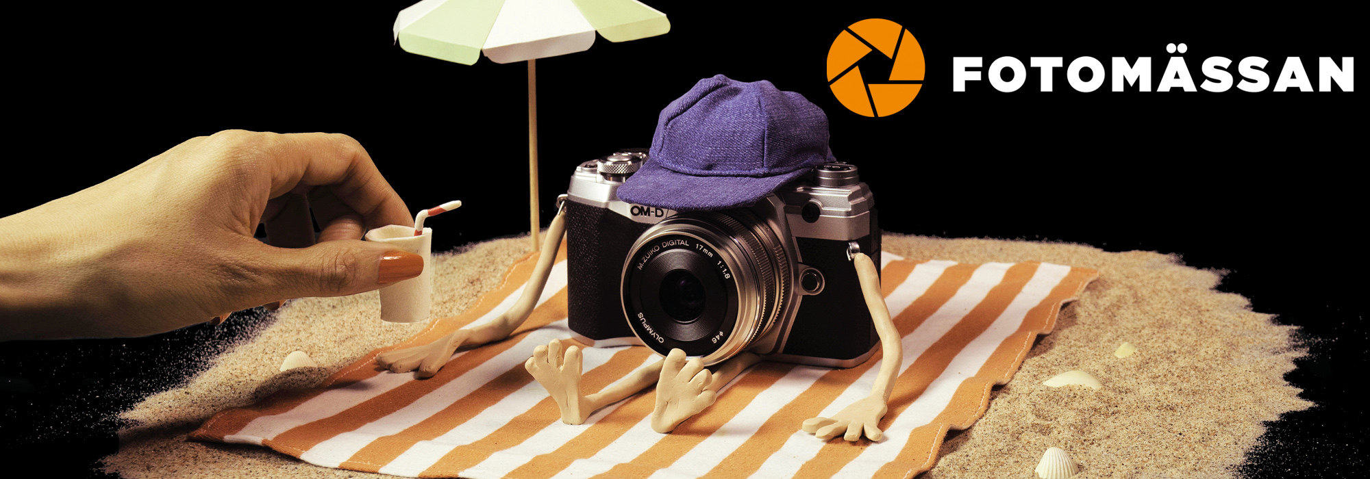 Fotomässan - en svart bakgrund med Fotomässans logotyp i vitt och med en orange illustrerad bländare. I samma bild syns en liten sandstrand och en kamera som är placerad på en filt intill ett parasoll. 